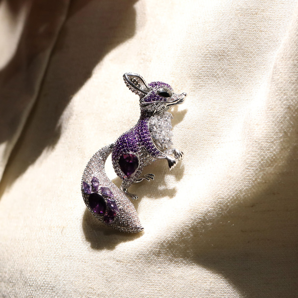 Purple and silver-tone fox brooch worn on a cream garment, shown in a close-up.