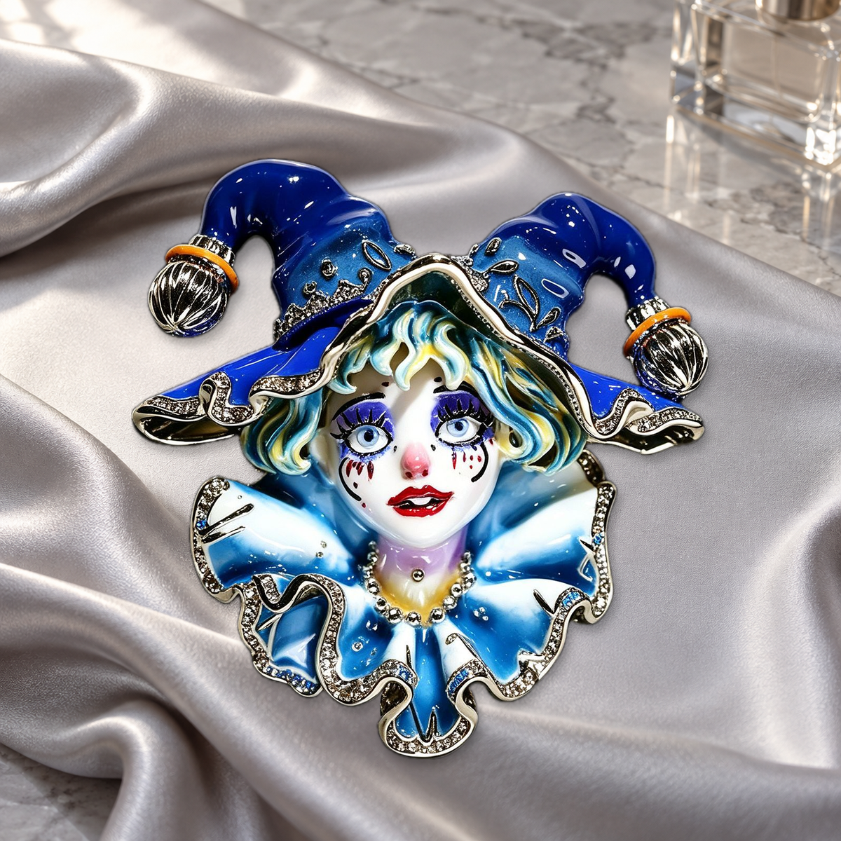 Blue jester brooch with a sculpted face, bell-tipped hat and ruffled collar on silver-gray satin on a dressing table.